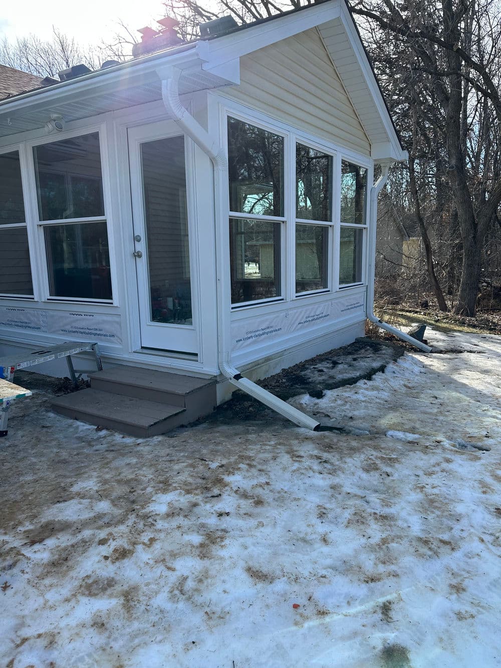 Sunlit corner of a house with large windows, surrounded by patchy snow and trees.
