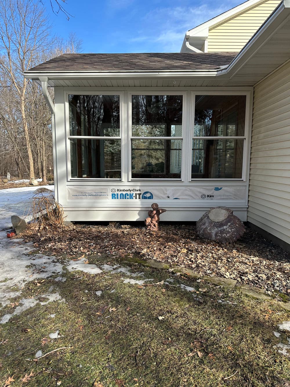 Sunroom exterior with branding, snow-covered ground, and stone features in a wooded area.