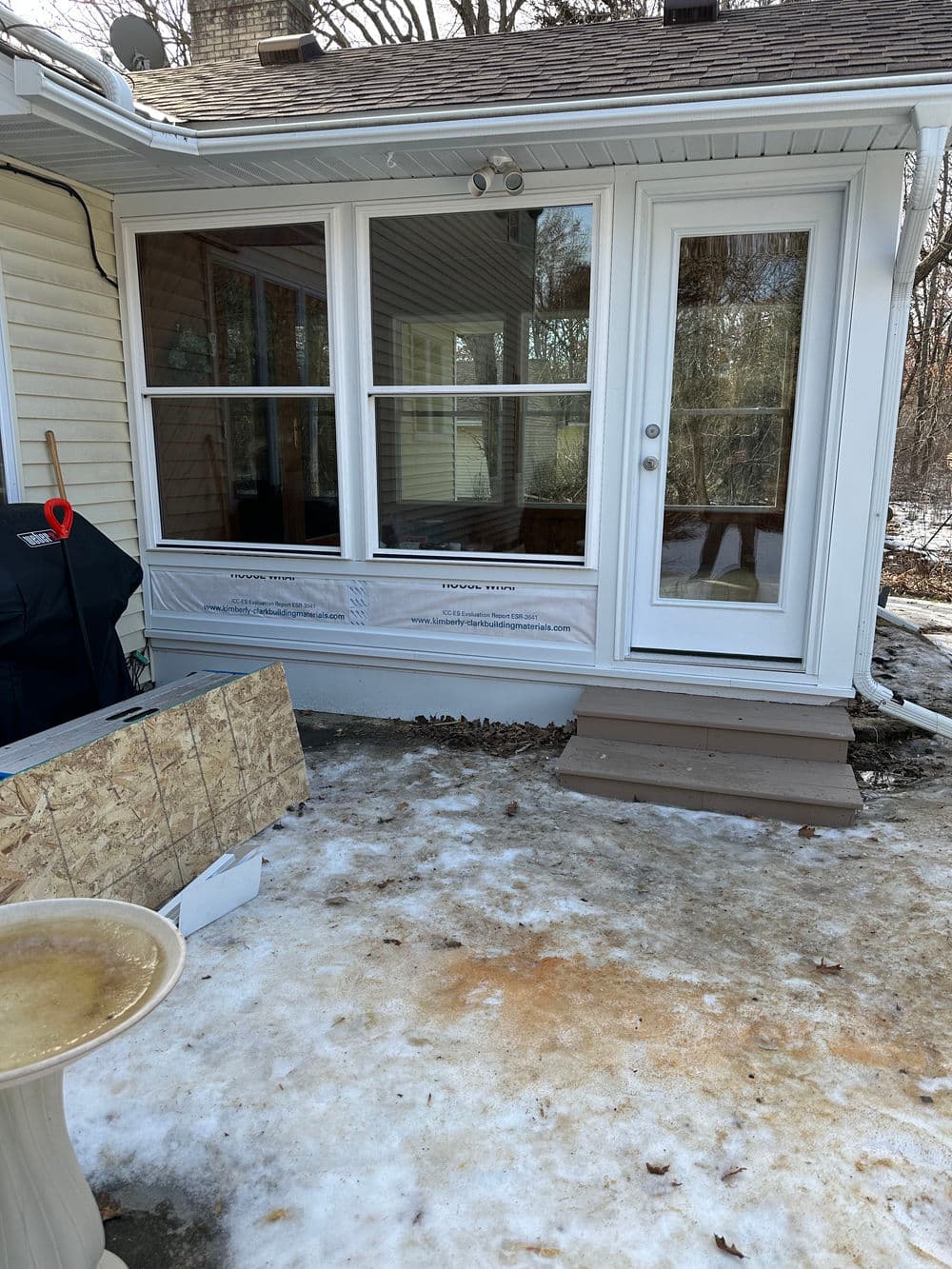Back porch area with large windows, steps, and construction materials on snowy ground.