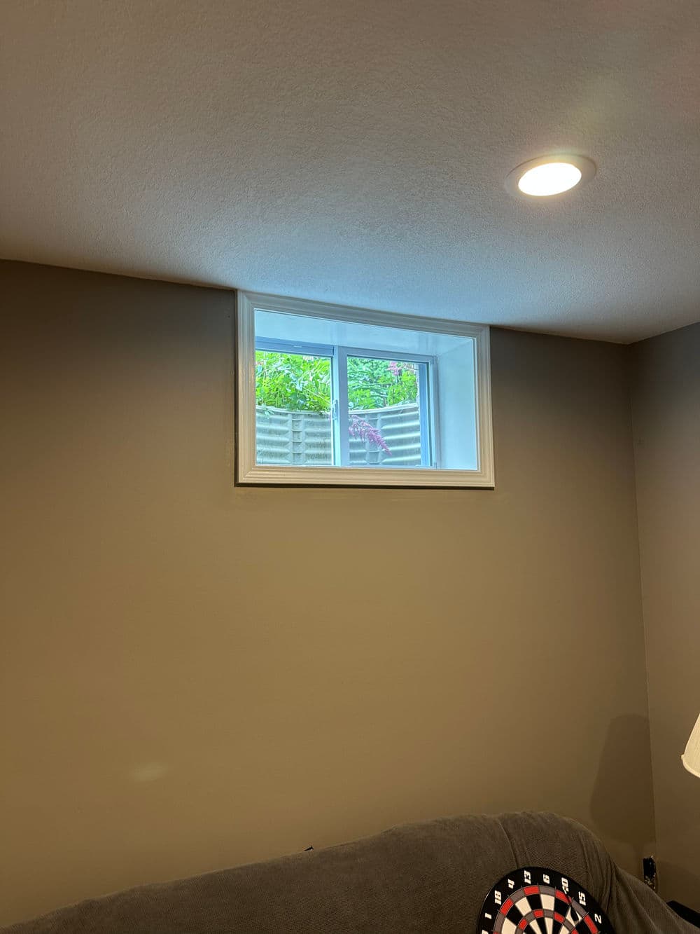 Bright basement window with a view of greenery, dartboard in the corner of the room.