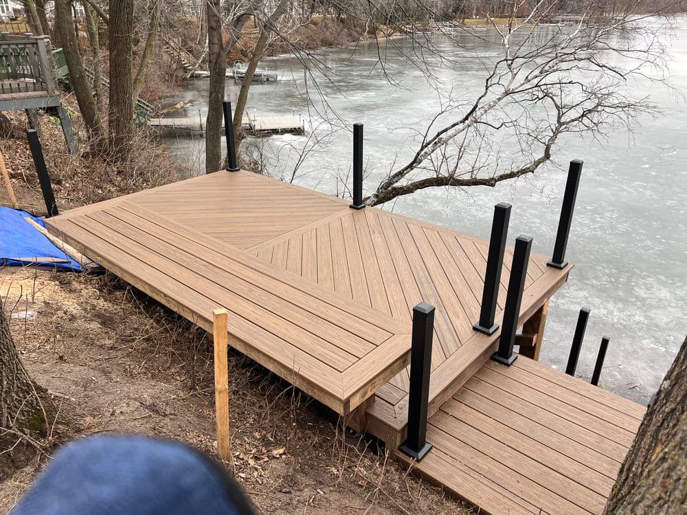 Newly constructed wooden dock by a frozen lake, featuring black posts and trees in the background.