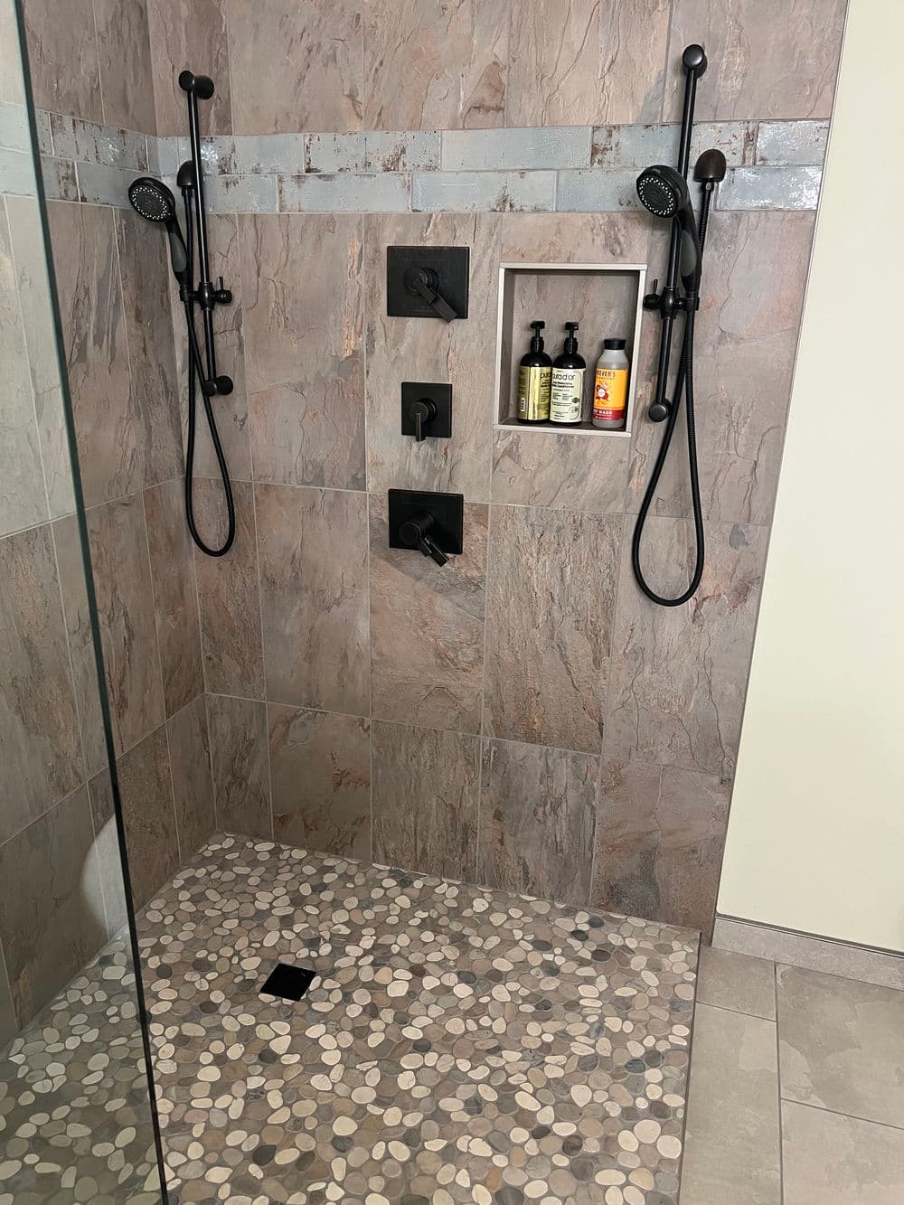 Modern shower design with dual black fixtures, stone tile walls, and pebble floor.