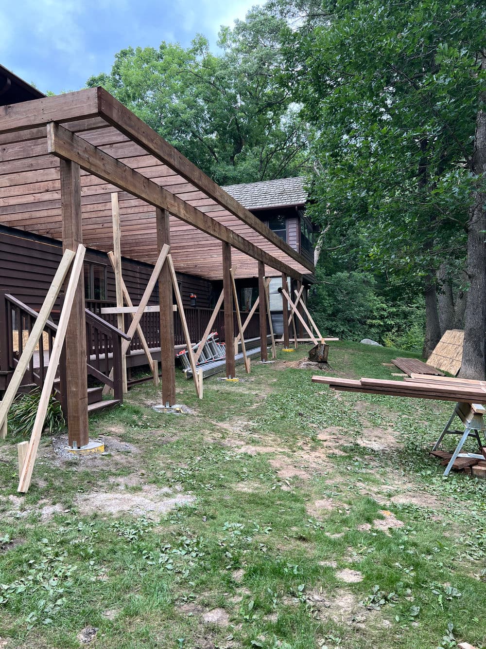 Construction of a wooden deck with supports beside a brown house in a wooded area.