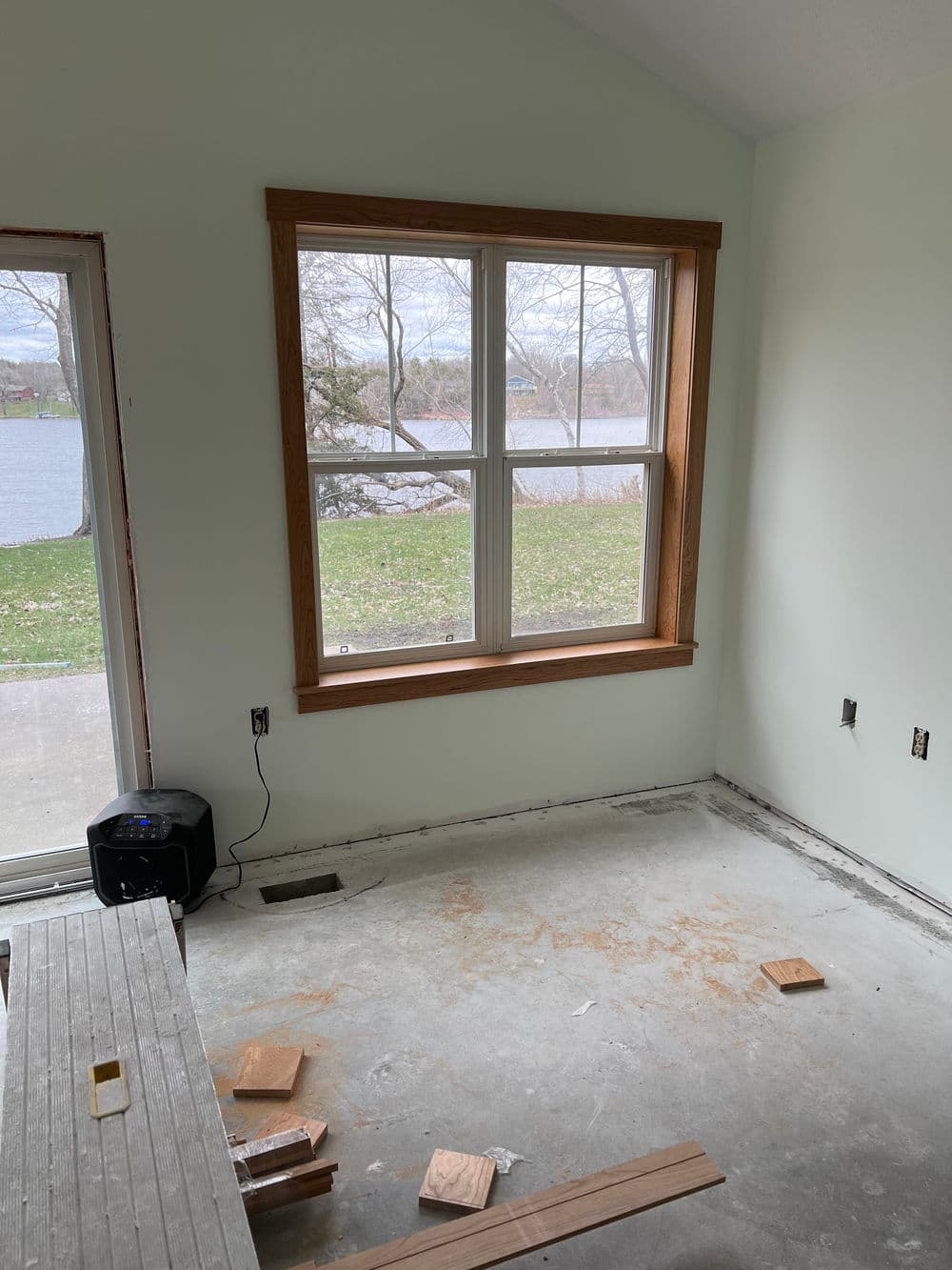 Room with large window overlooking a lake, unfinished floor, and construction materials.