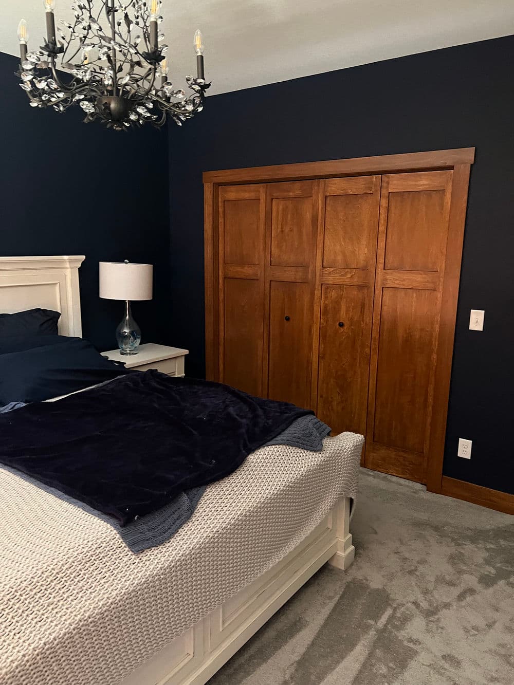Cozy bedroom with dark blue walls, wooden closet doors, and elegant chandelier lighting.
