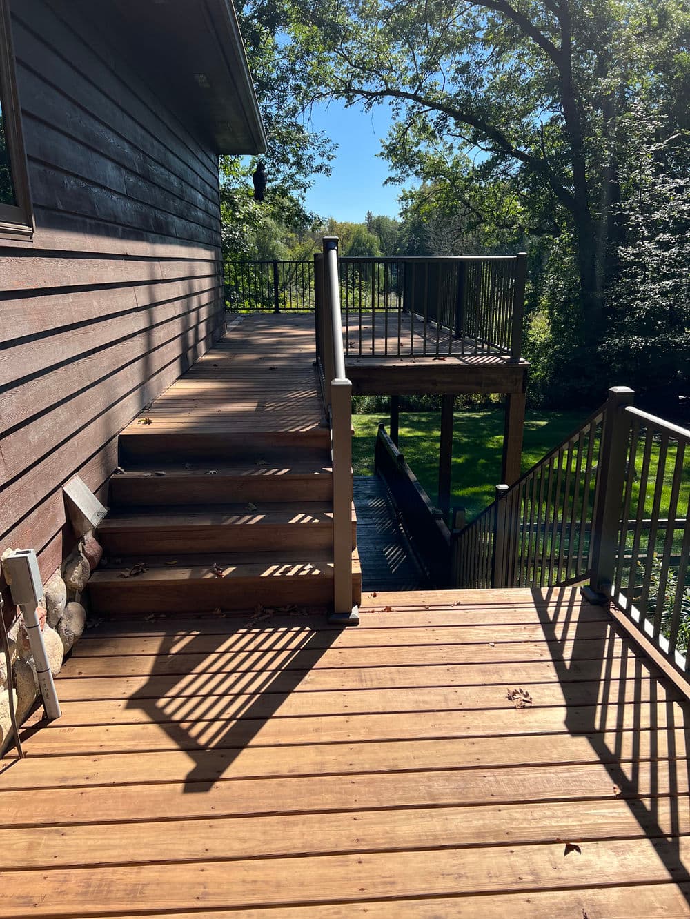 Wooden deck with stairs, surrounded by trees and sunlight, showcasing outdoor relaxation space.