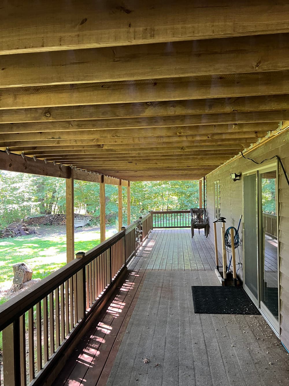 Covered porch with wooden beams and railing, overlooking a green yard and trees.
