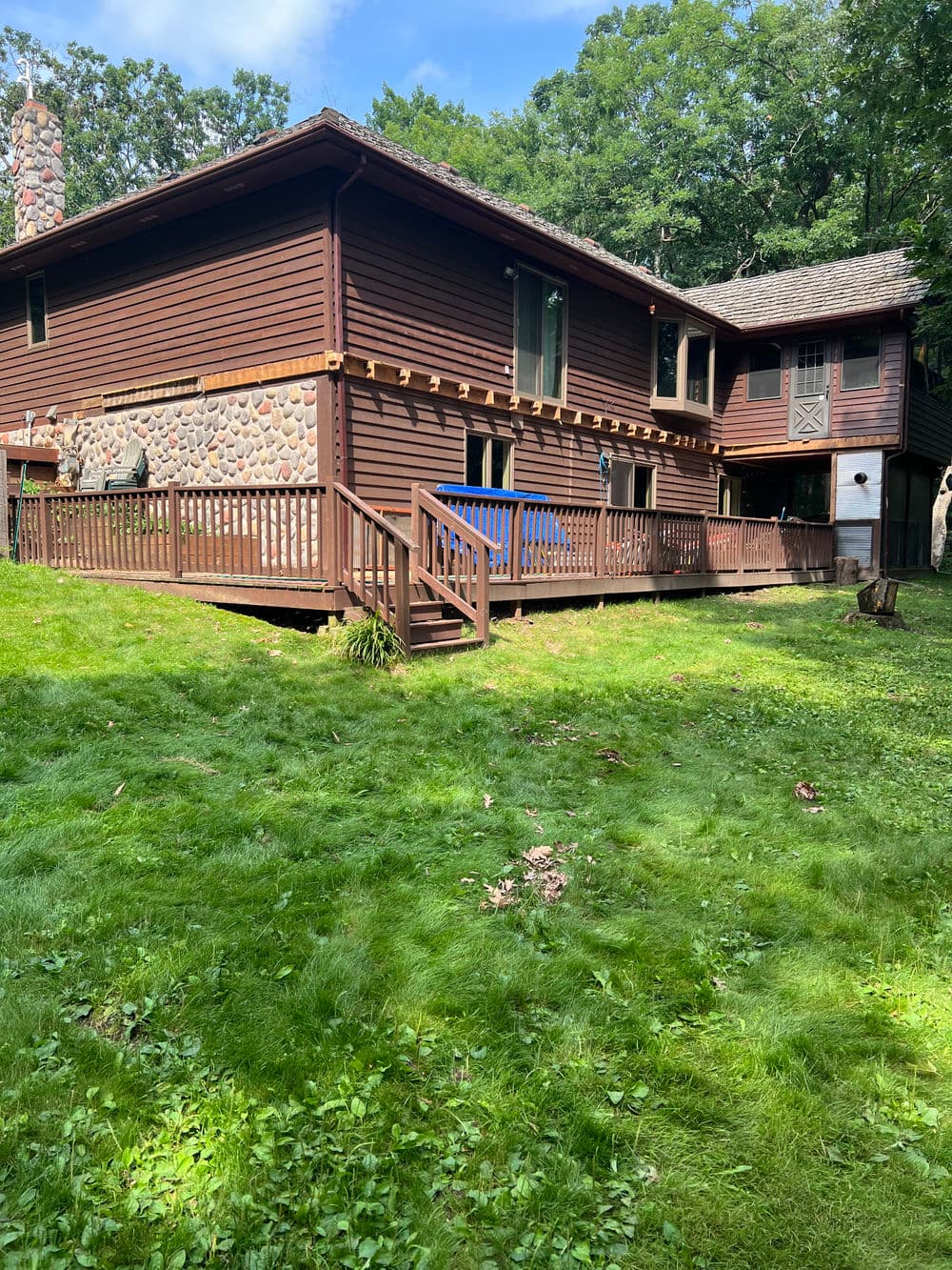 Wooden house with stone accents and a deck, surrounded by green lawn and trees.