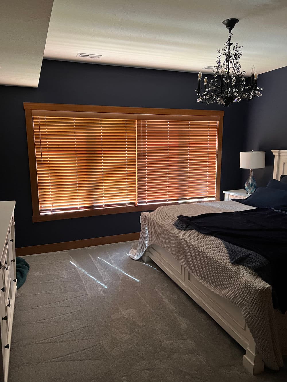Cozy bedroom with dark walls, wooden blinds, a white bed, and stylish chandelier lighting.