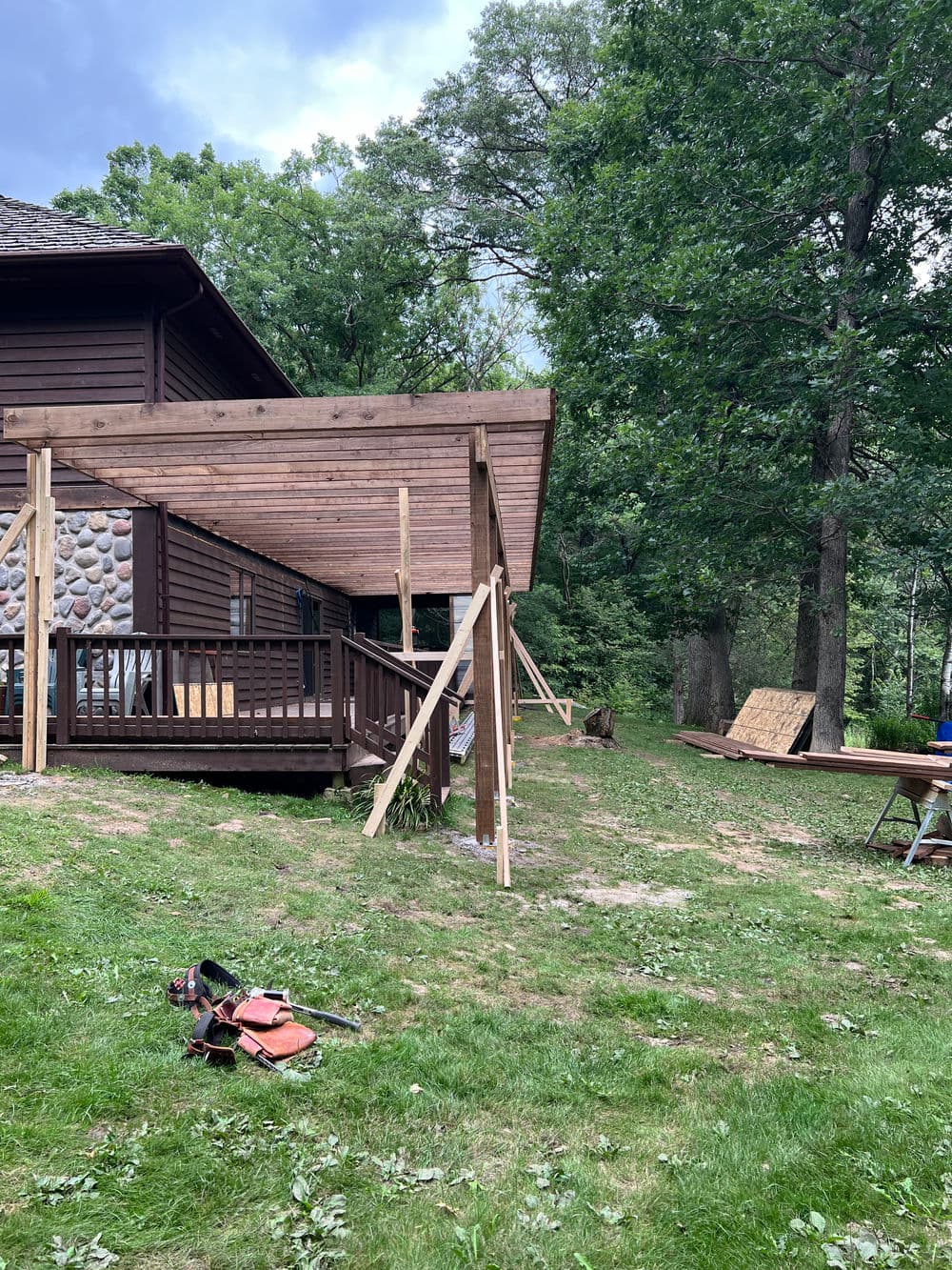 Wooden deck under construction beside a brown house, surrounded by green trees and grass.