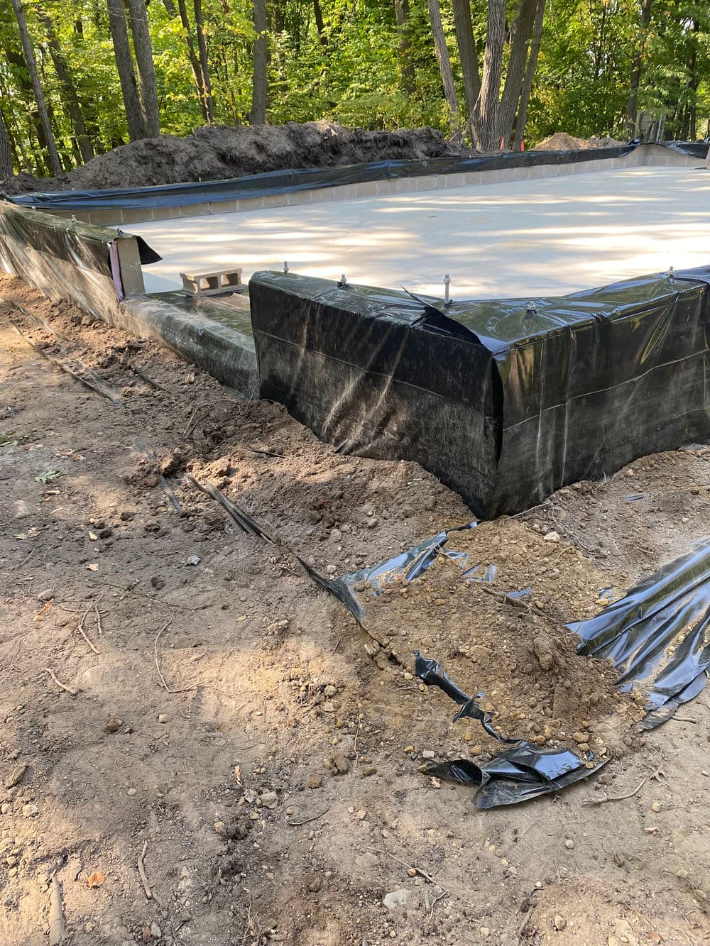 Construction foundation site with gravel and plastic sheeting surrounded by trees.