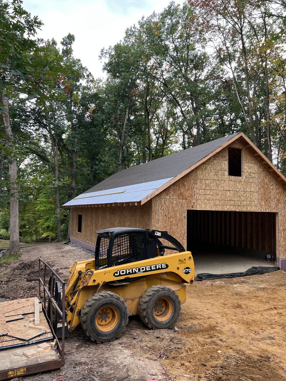 Construction site featuring a John Deere skid steer near a wooden-framed house in a forest.