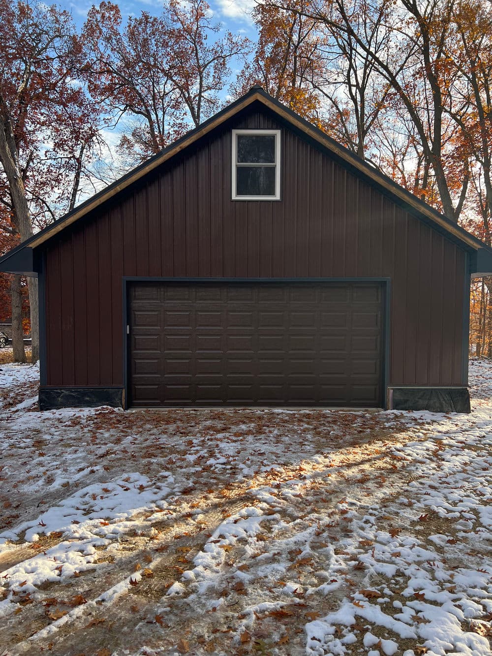 Brown garage with large door, surrounded by autumn leaves and trees in a snowy landscape.