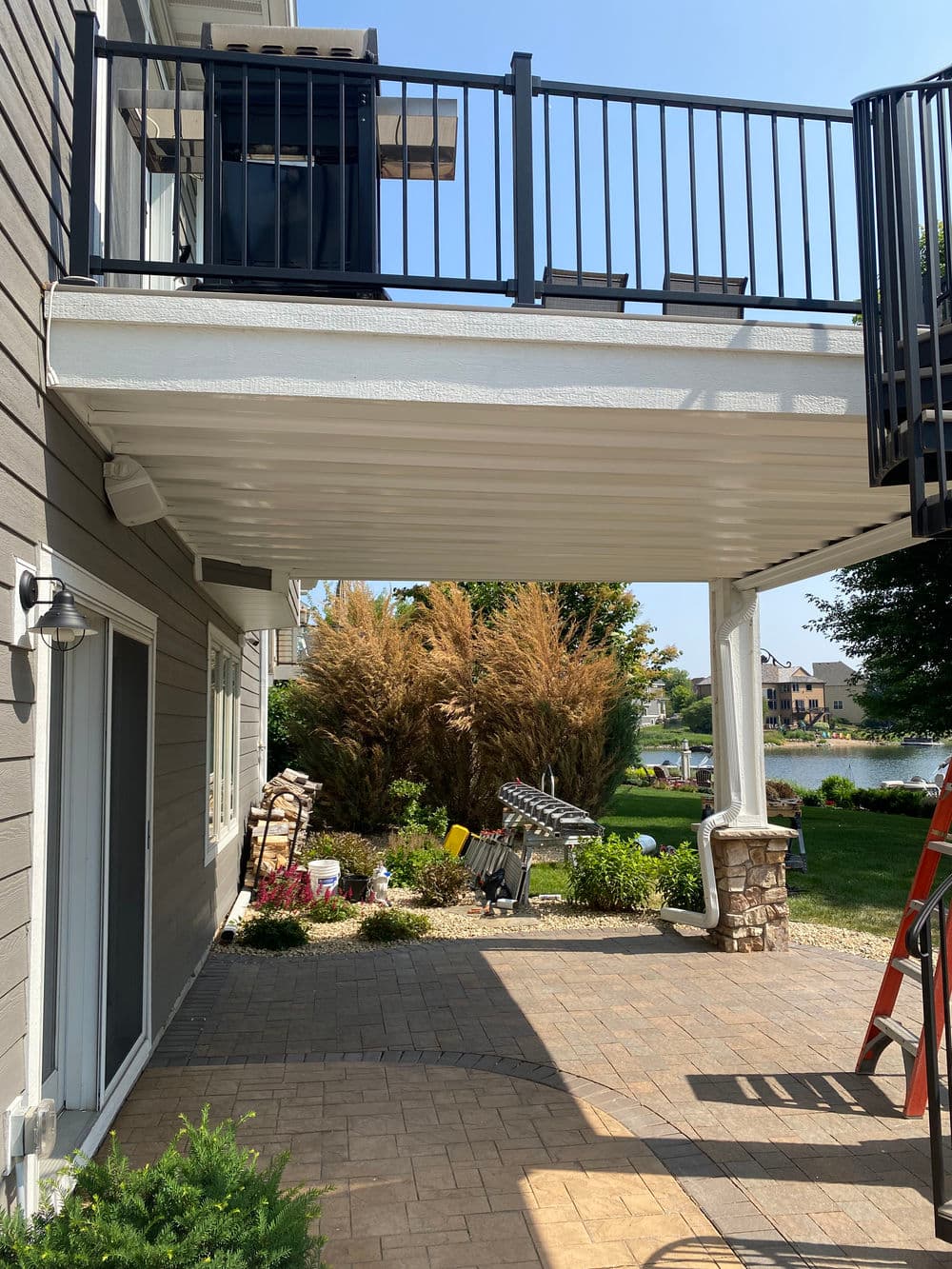 Under-deck patio area with pavers, landscaping, and waterfront view, featuring a wooden railing.