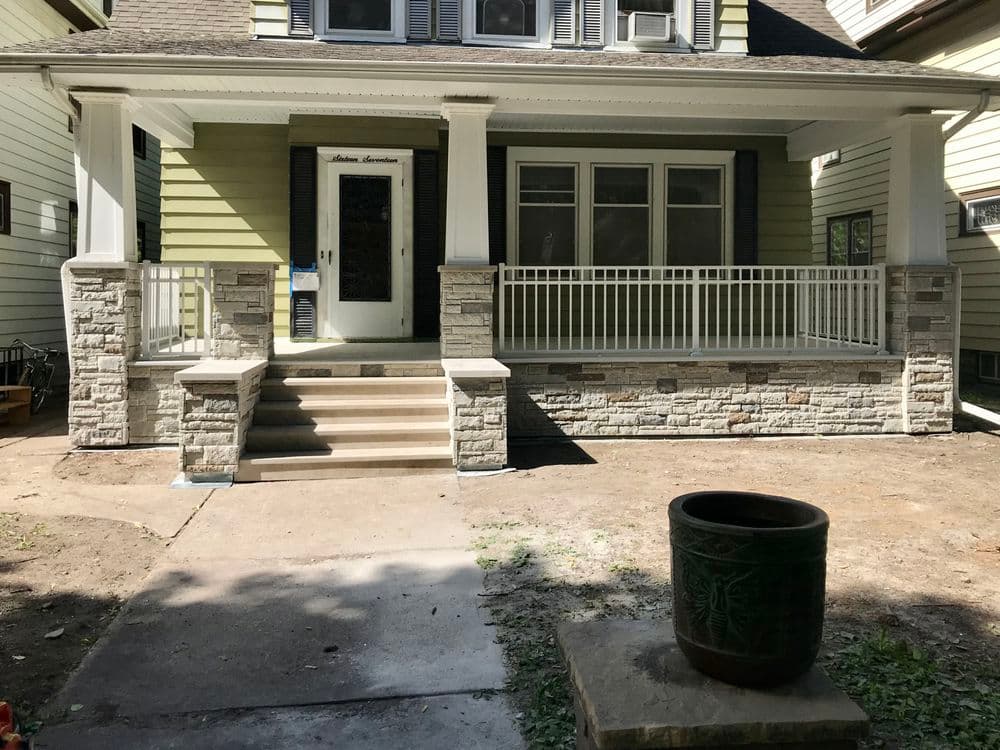 Renovated home entrance with stone columns, porch, and potted plant in yard.