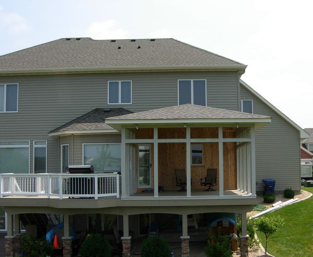 Modern home exterior featuring a deck and screened porch with two chairs. Green landscaping.