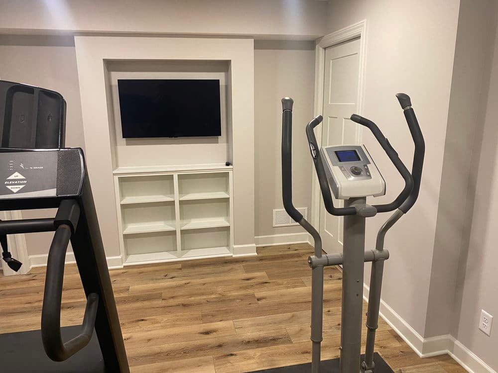 Home gym with elliptical machine, treadmill, and modern decor. TV and shelving unit in background.