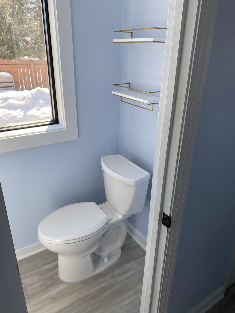 Modern bathroom with a toilet, light blue walls, and minimal shelving above.