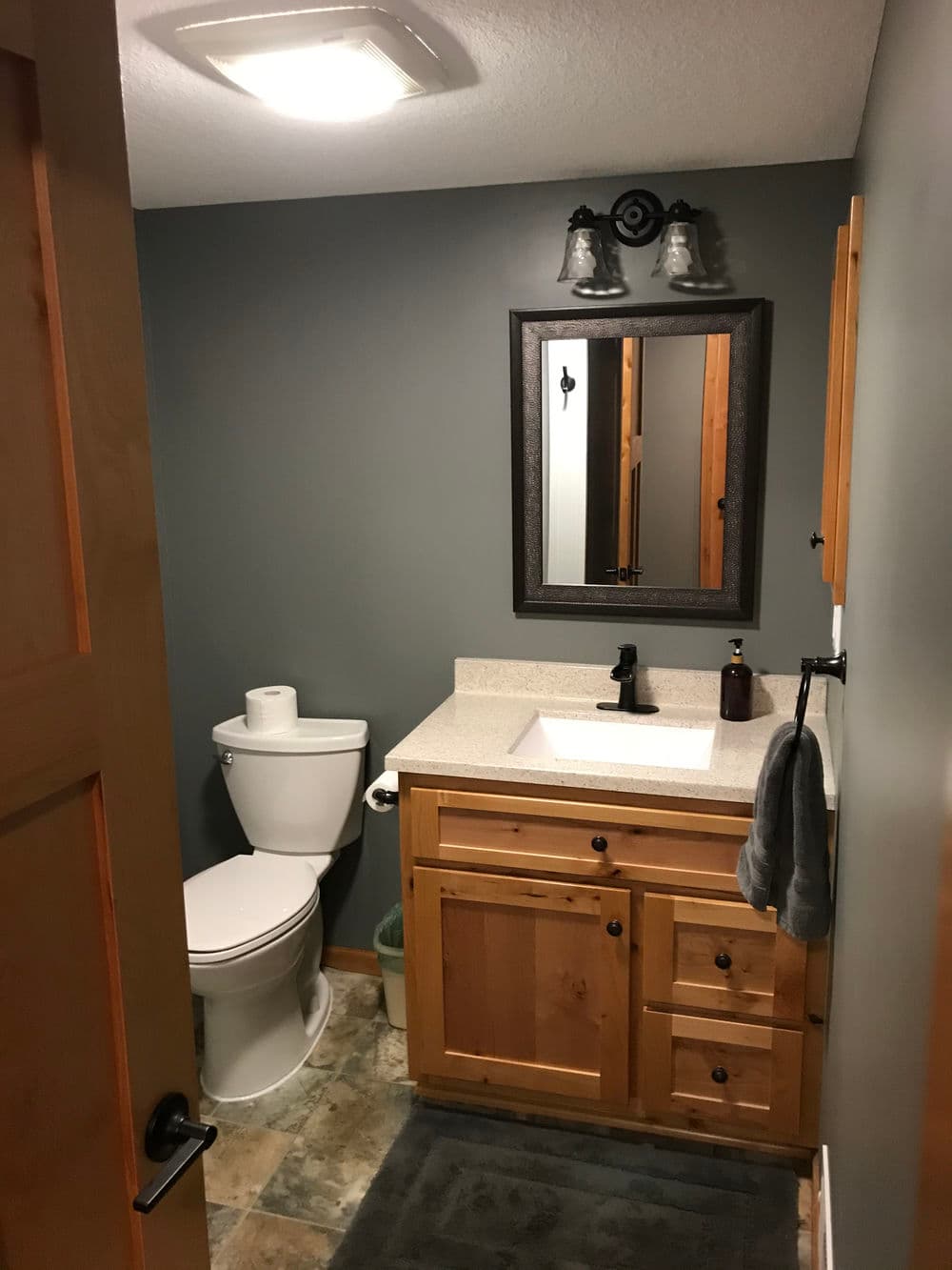 Modern bathroom with wooden cabinetry, mirror, and stone flooring. Toilet and sink visible.